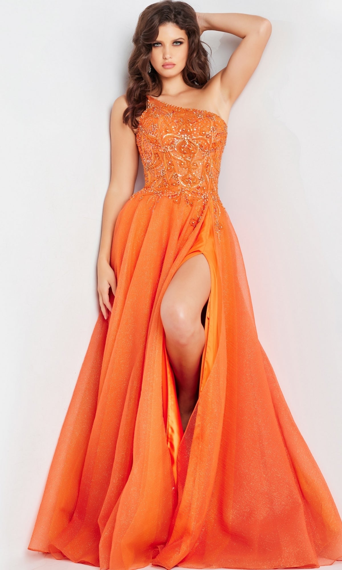 A woman poses against a white background wearing the Jovani One-Shoulder Long Prom Ball Gown 25688 in orange, featuring embellishments and a high slit.